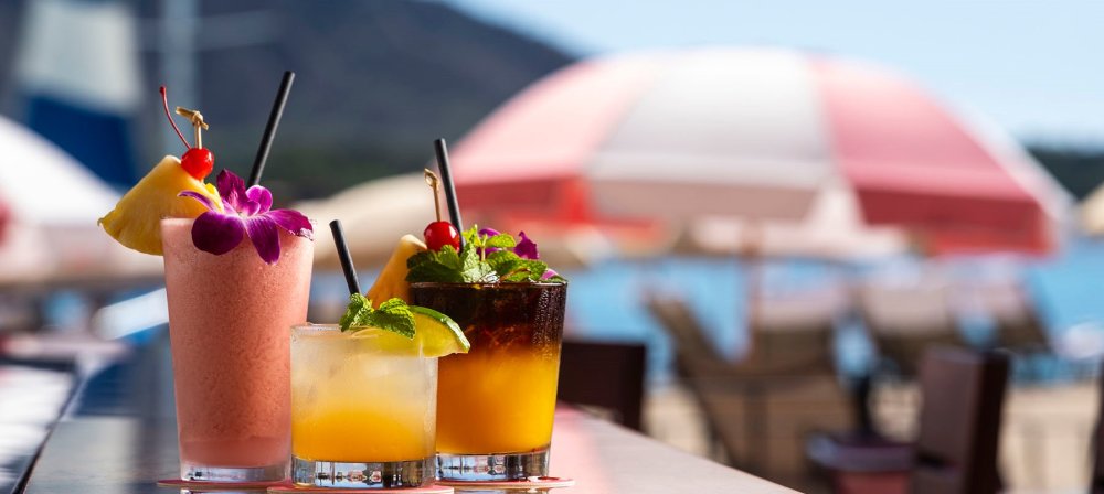 Tropical drinks with straws and garnishes on a bar, beach and umbrellas in background.