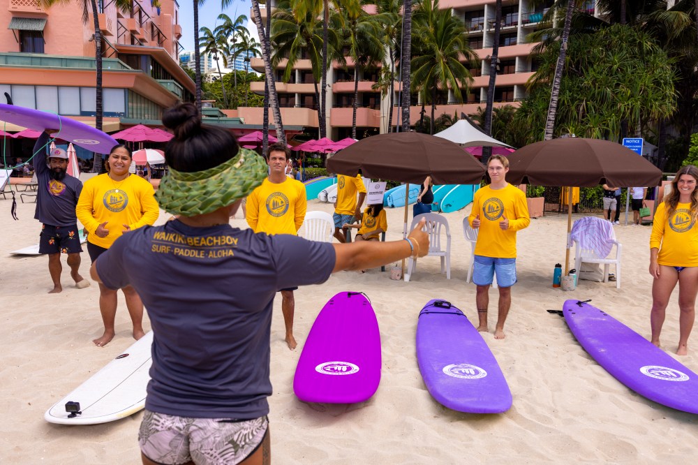 Surf instructor giving a lesson on the beach with students in yellow shirts holding surfboards.