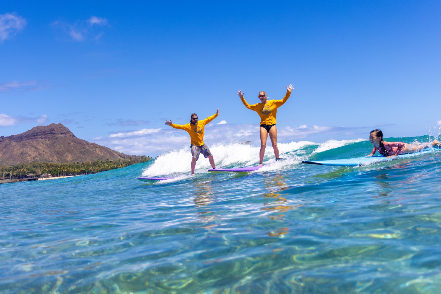 Three surfers in bright clothes catching waves, with a mountain and clear sky in the background.