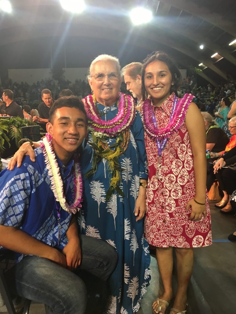 Three people smiling, wearing floral lei necklaces in a crowded hall.