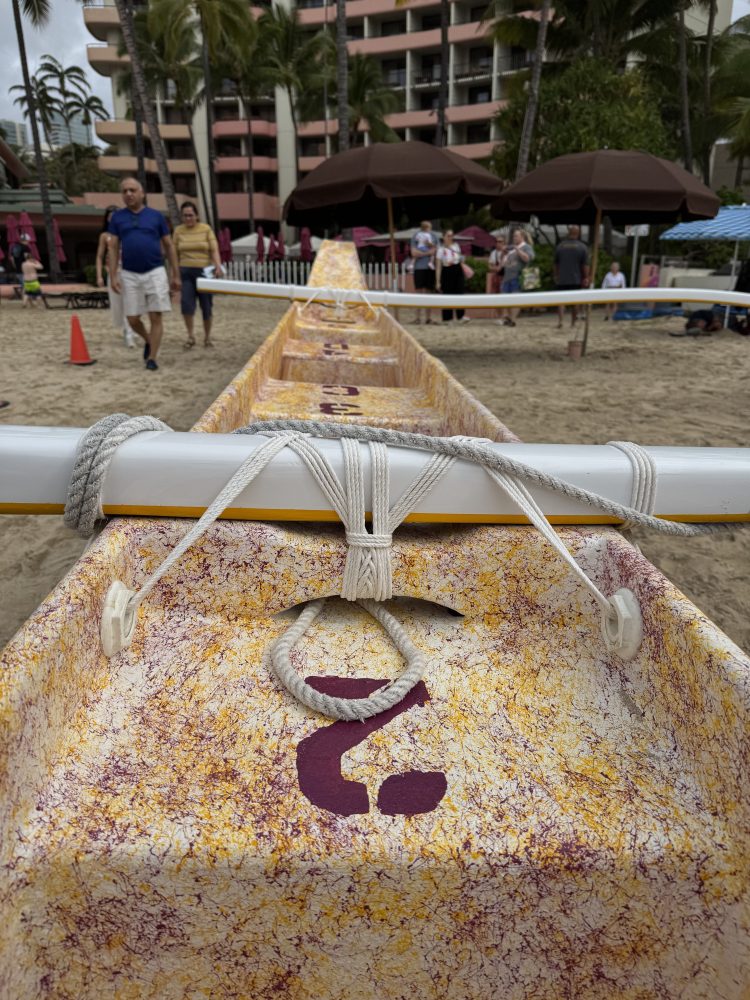 Close-up of a canoe with a yellow and white pattern on a sandy beach, lined with palm trees in the background.