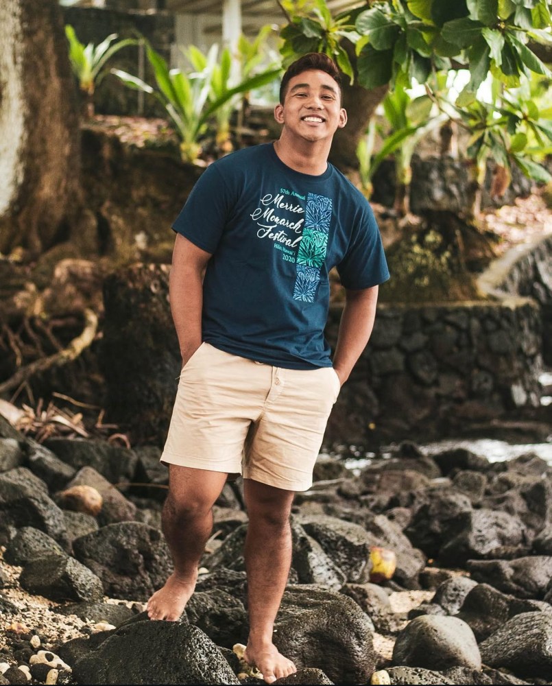 Person in blue shirt and shorts smiles, standing barefoot on rocky shore beside greenery.