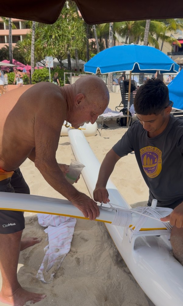 Two men repairing a surfboard on a beach under umbrellas.