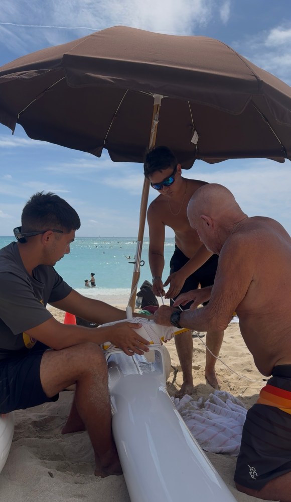 Three people setting up beach equipment under an umbrella by the sea.