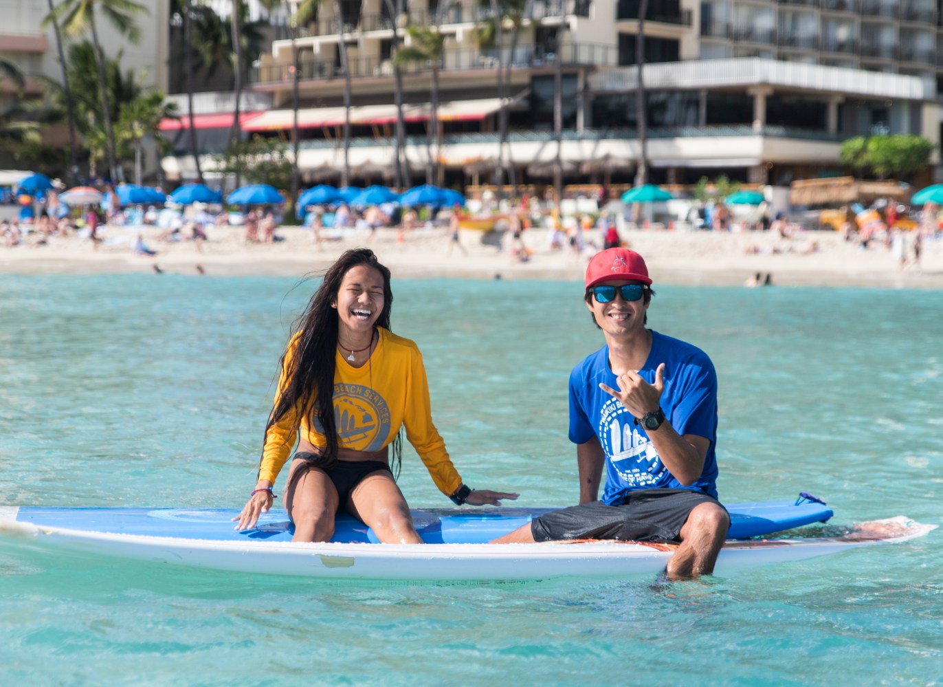 Two people sitting on a surfboard in the ocean near a busy beach.