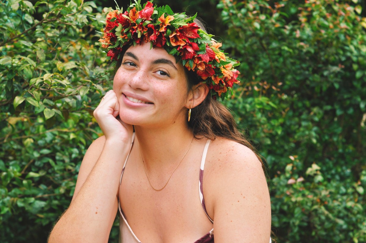 Smiling person with floral crown, leaning on hand, surrounded by greenery.