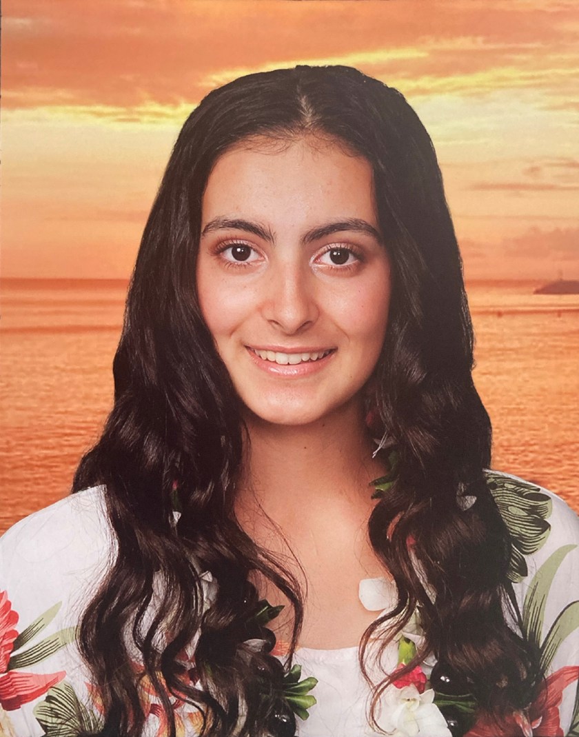 Smiling person with long hair, wearing a floral outfit against a sunset over the ocean.