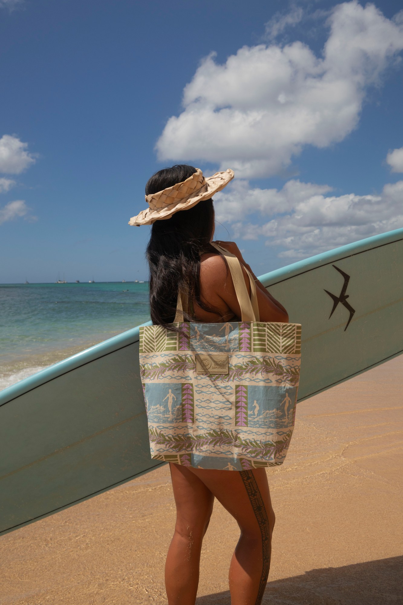 Person with surfboard on beach, wearing a sunhat and patterned tote.