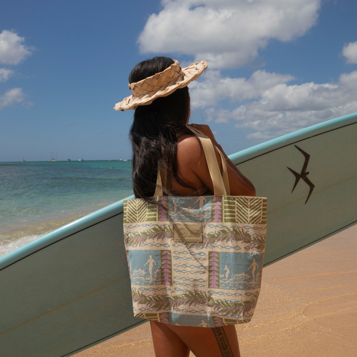 Person with surfboard on beach, wearing a sunhat and patterned tote.