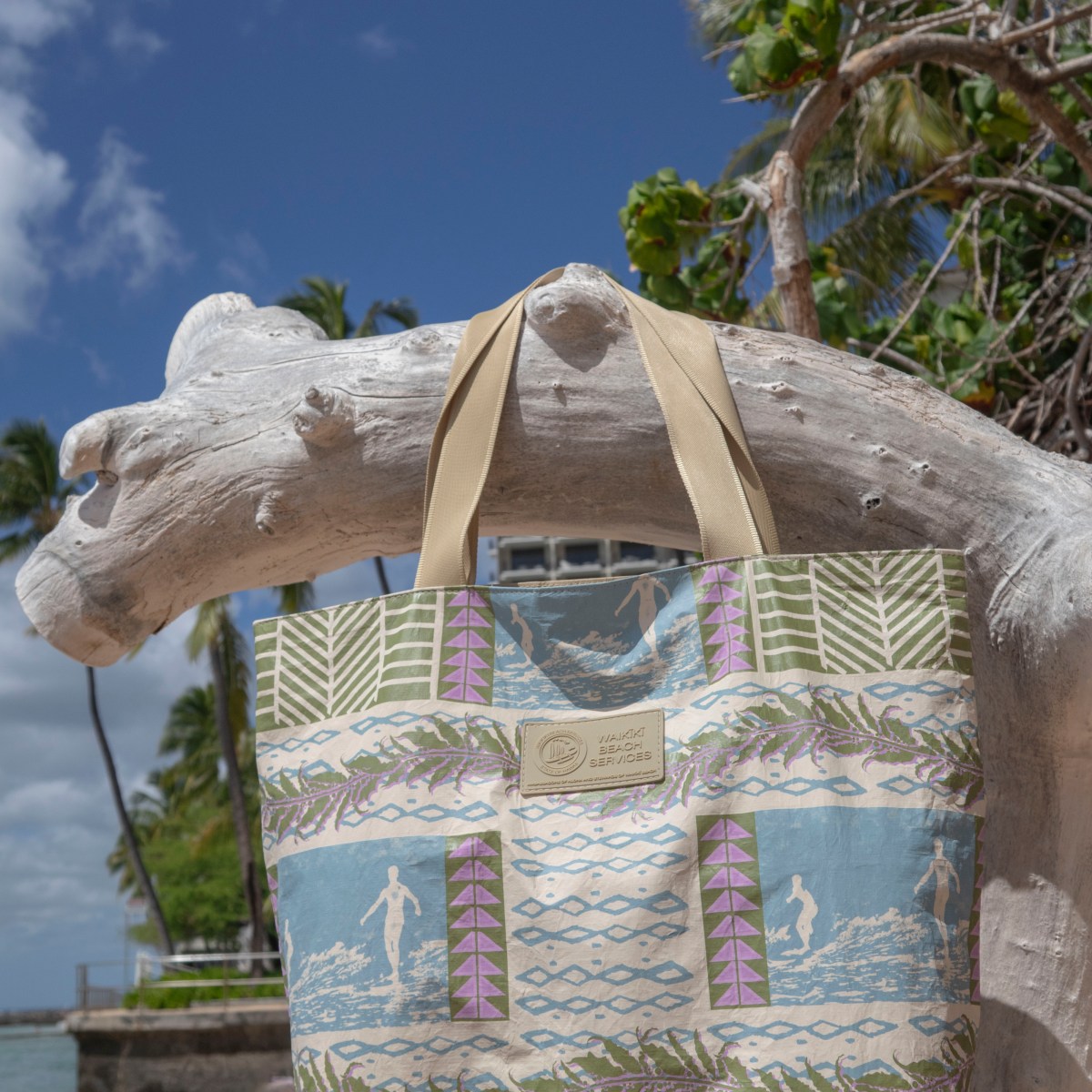 Tote bag hanging on driftwood at a beach with palm trees and blue sky.