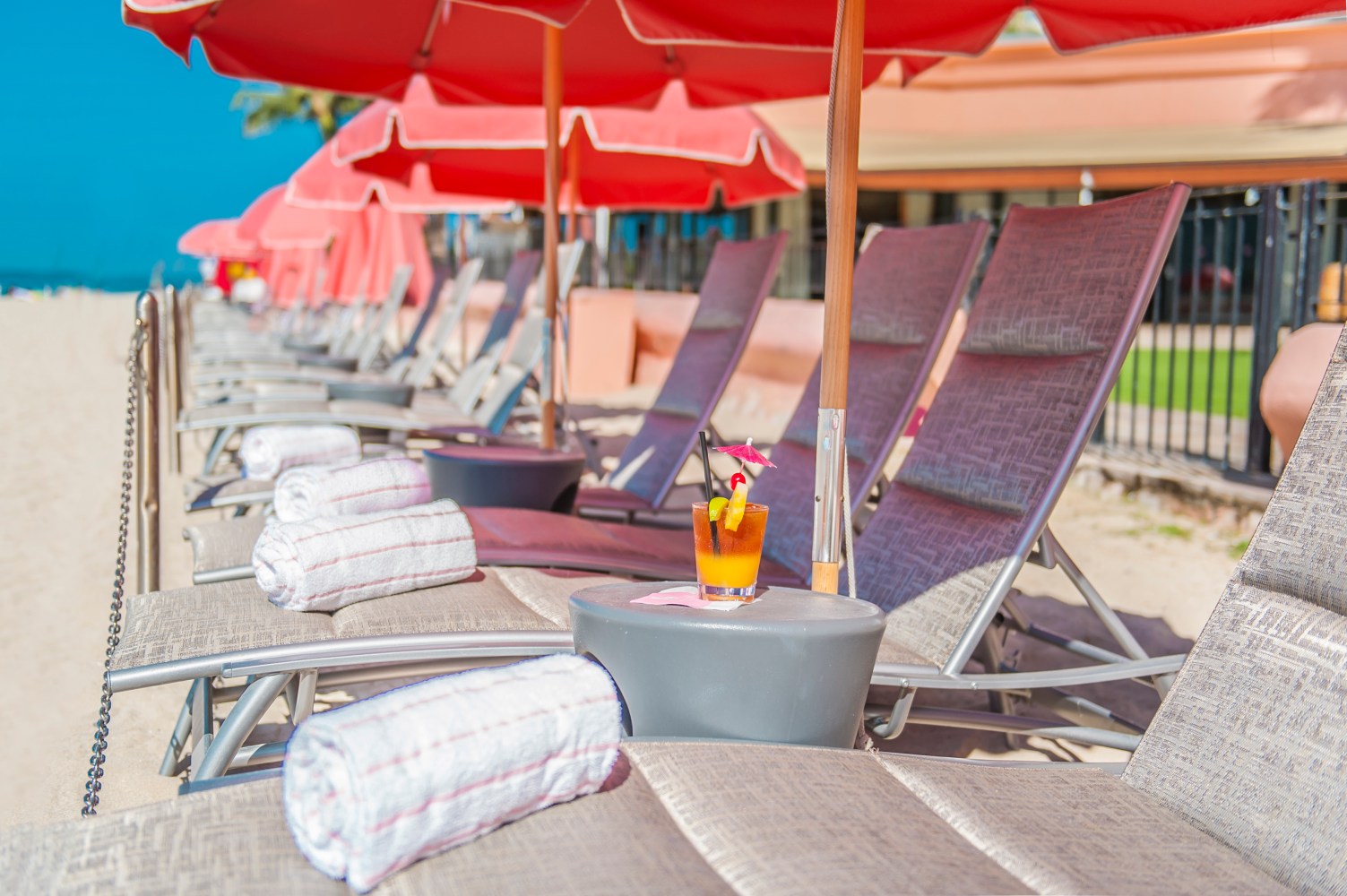 Beach chairs with towels and drinks under red umbrellas on a sandy beach.
