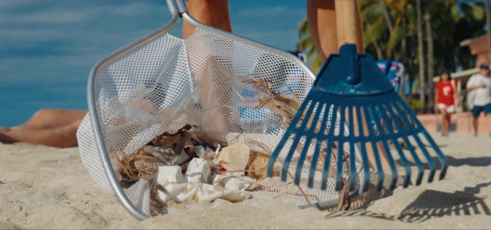 Person cleaning beach, collecting trash with a rake and net.