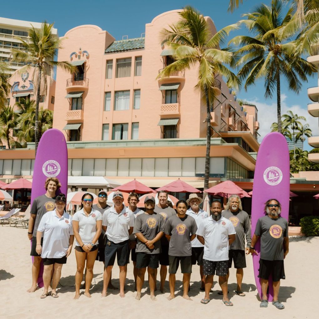 Group of people on a sandy beach with two purple surfboards, palm trees, and a peach building in the background.