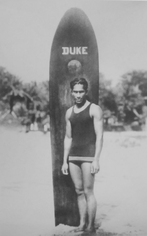 Man in tank top with surfboard labeled 'Duke' on a beach.