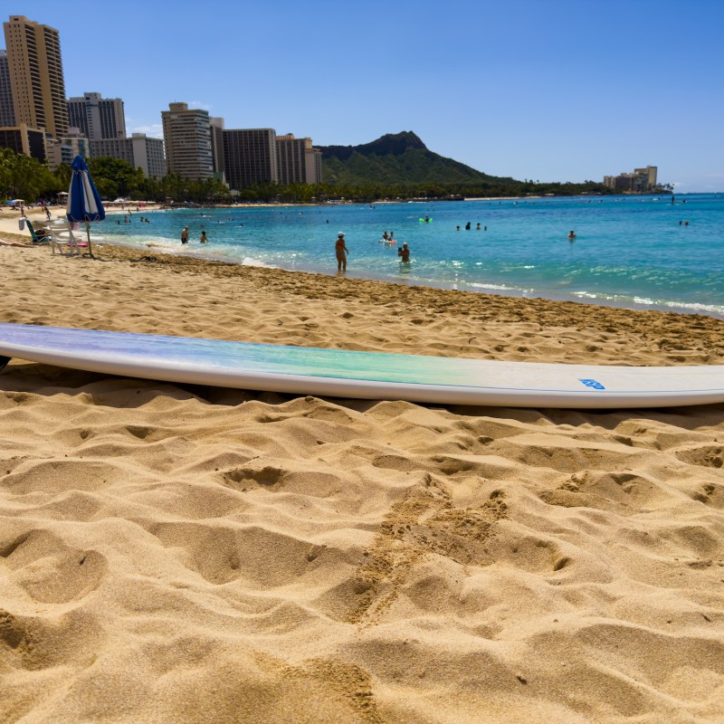 Surfboard on sandy beach with ocean and city skyline in the background.