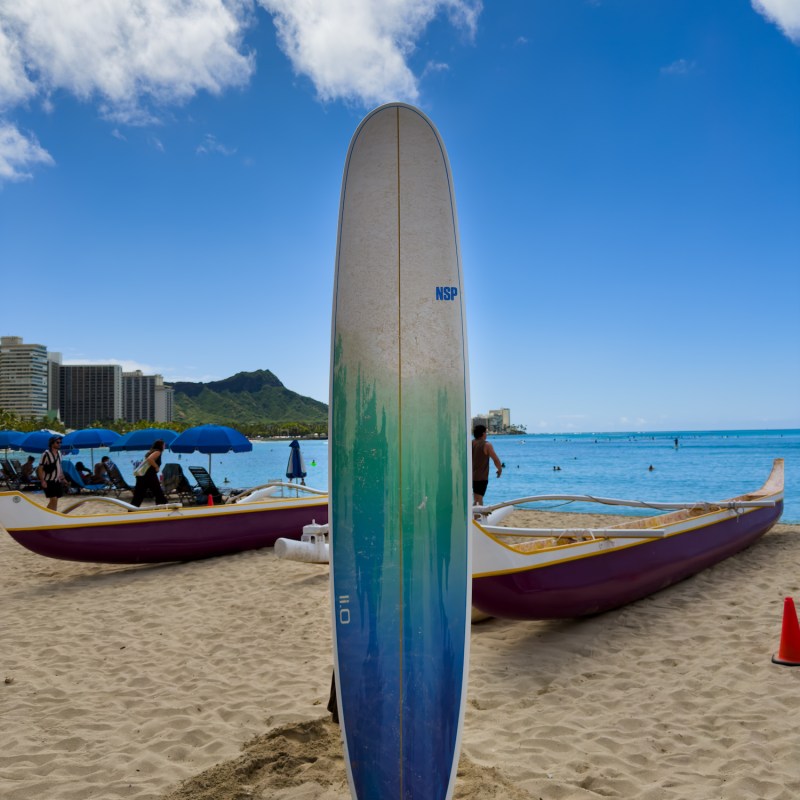 Colorful surfboard standing on a beach with boats and ocean in the background.