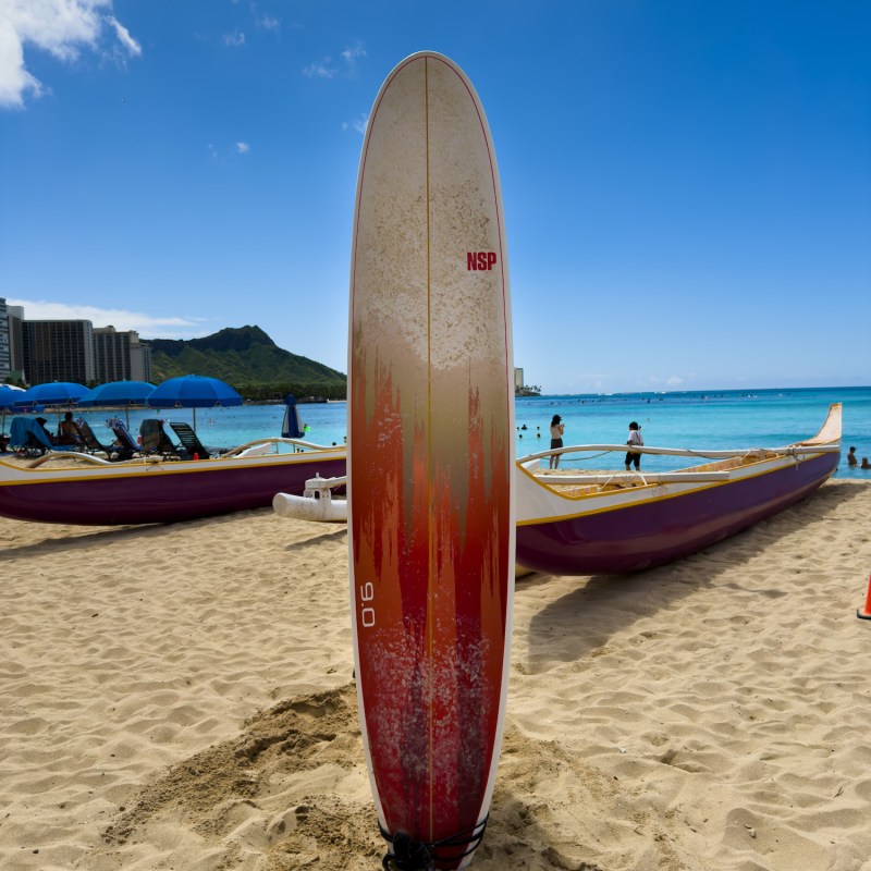 Surfboard standing upright on sandy beach with ocean and canoes in background.