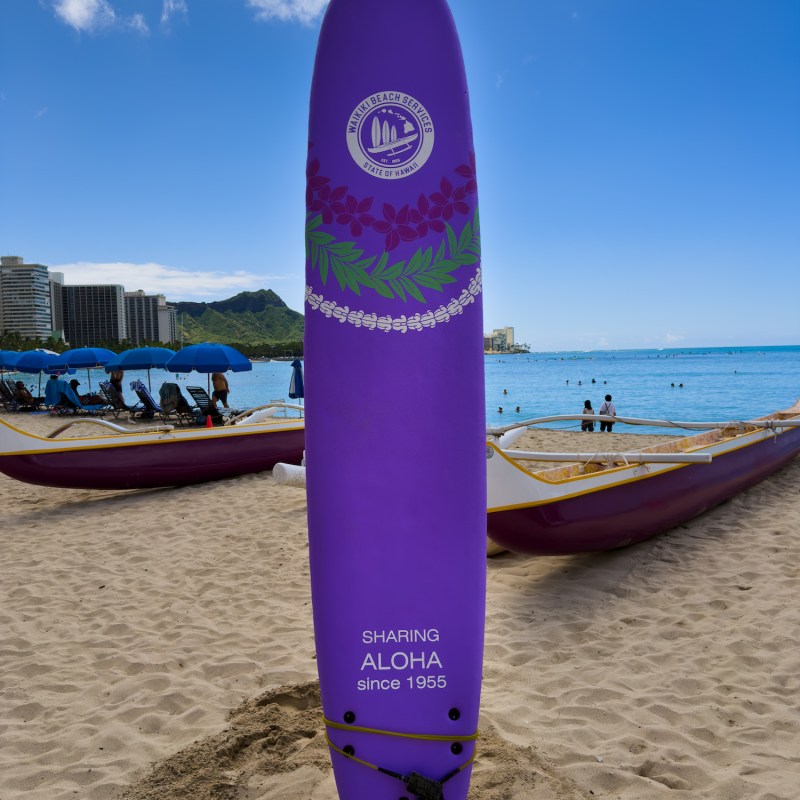 Purple surfboard with 'Sharing Aloha' text standing on beach sand.
