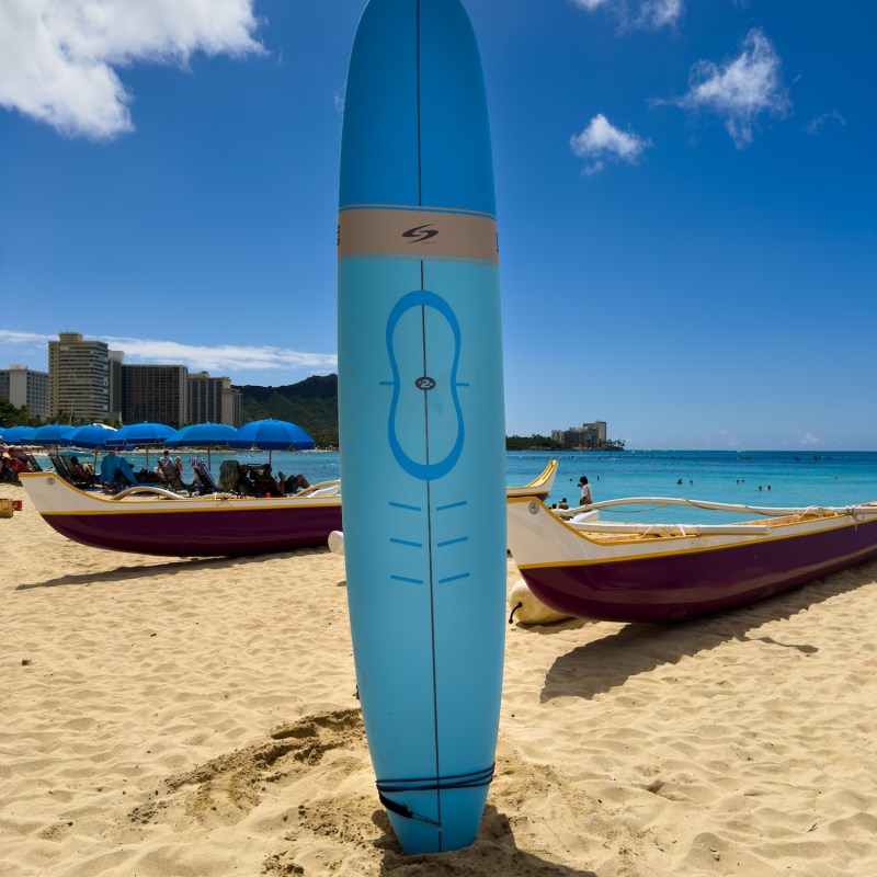 Blue surfboard standing upright on sandy beach, with ocean and canoes.