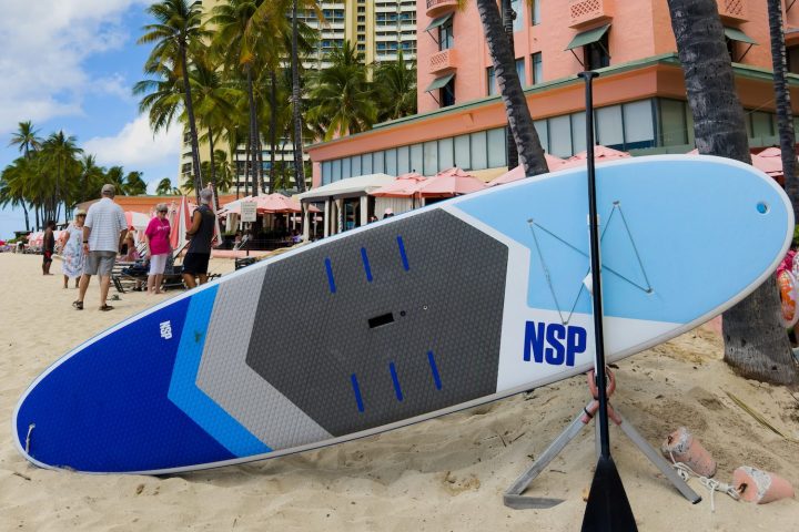 Surfboard and palm trees on a sandy beach with tall hotels in the background.