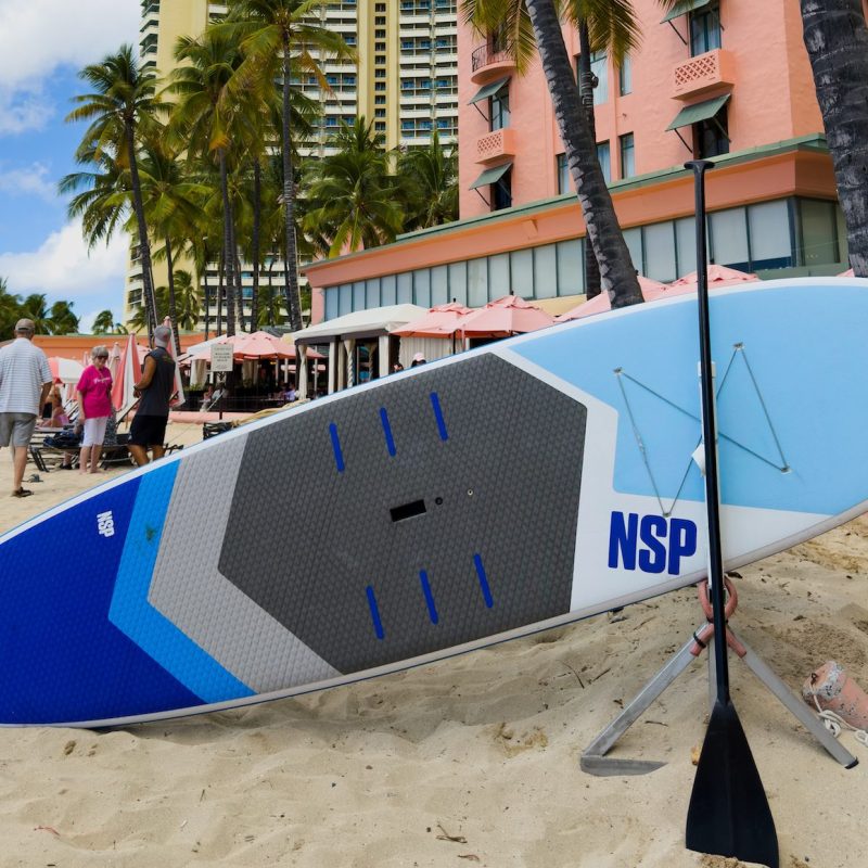 Surfboard and palm trees on a sandy beach with tall hotels in the background.