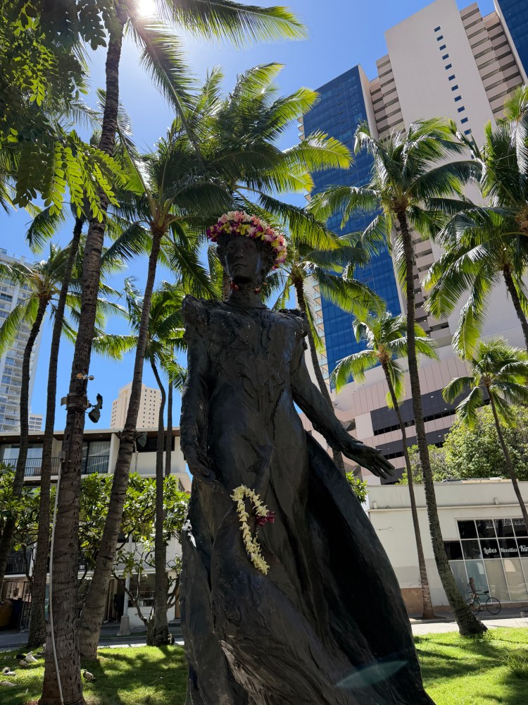 Statue with floral crown and lei, surrounded by palm trees and tall buildings.